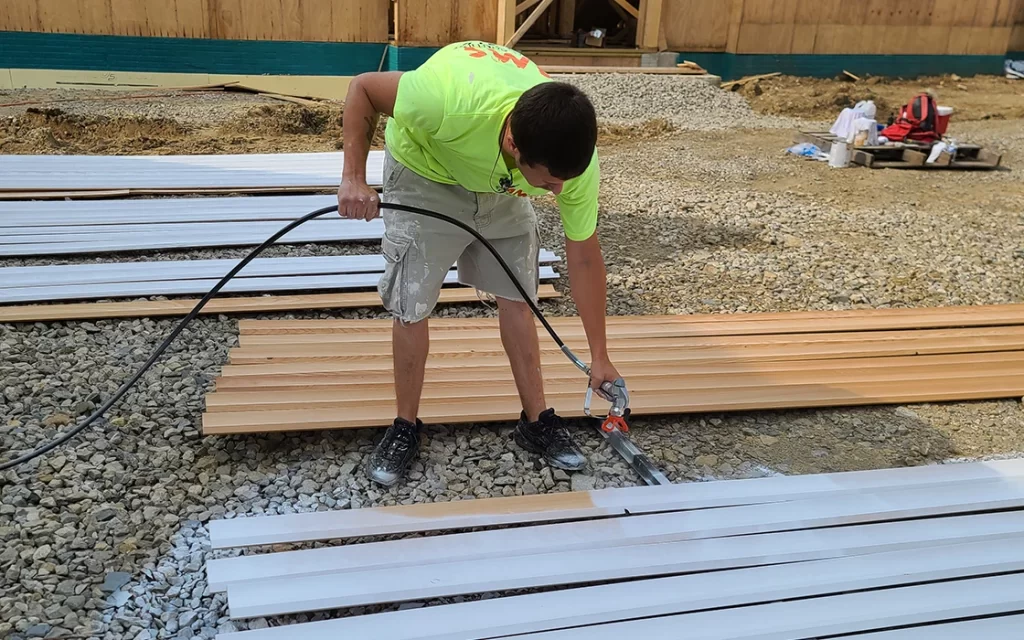 Construction worker in a neon shirt cutting a metal strip with a power saw on a lumber-strewn construction site.